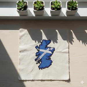May include: A blue embroidered patch in the shape of Scotland with a white and blue cross design, placed on a square of off-white fabric with frayed edges. Four small square white pots with green succulents are arranged above.