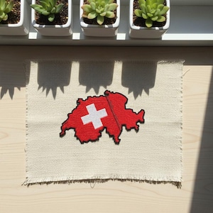 May include: A red and white embroidered patch in the shape of Switzerland, with a white cross on a red background. The patch is on a beige, fringed fabric square. The image is taken in natural light.