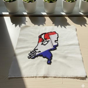 Op de afbeelding: Geborduurde patch in de vorm van Nederland, met de kleuren van de Nederlandse vlag: rood, wit en blauw. De patch is geplaatst op een stuk ecru stof met een gerafelde rand. Op de achtergrond staan kleine vetplanten in potten.