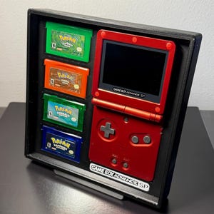 以下が含まれることがあります： 黒いディスプレイケースに入った、赤いゲームボーイアドバンスSP本体と4つのポケモンゲームカートリッジ。カートリッジは緑、オレンジ、緑、青です。コンソールには「GAME BOY ADVANCE SP」の文字があります。