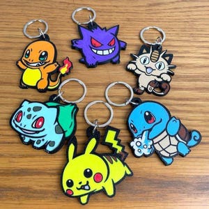 May include: Seven cartoon character keyrings in various colours, including orange, purple, blue, green, and yellow. Each keyring features a different character design and has a silver metal ring attached for keys.