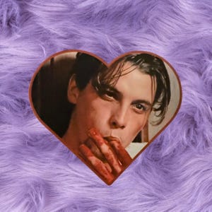 May include: Heart-shaped sticker featuring a portrait of a man with blood on his fingers and mouth. The sticker has a brown border and is set against a purple, fluffy background. The man has dark hair and is looking at the viewer.