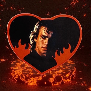 May include: A heart-shaped patch featuring a portrait of a man with long hair, set against a black background. The patch has a red border with flame-like designs. The patch is placed on a fiery, lava-like pedestal.