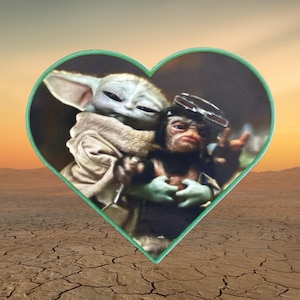 May include: A heart-shaped pin featuring a printed image of a green, large-eared creature wearing a tan robe embracing a smaller, dark-furred creature with goggles on its head. The pin has a thin, light green border and a smooth, glossy finish.