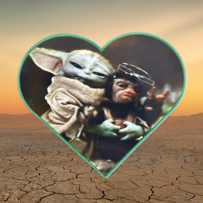 May include: A heart-shaped pin featuring a printed image of a green, large-eared creature wearing a tan robe embracing a smaller, dark-furred creature with goggles on its head. The pin has a thin, light green border and a smooth, glossy finish.