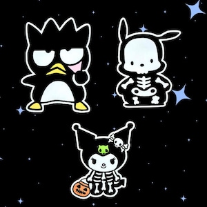May include: Three cartoon character stickers on a black background with blue stars. One is a black penguin with yellow feet, one is a white skeleton figure with long ears, and one is a black character wearing a skeleton costume holding a pumpkin.