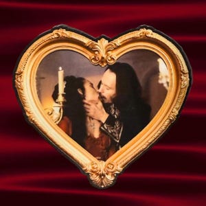 May include: A heart-shaped picture frame with a gold ornate design. The frame holds a sepia-toned image of a couple kissing. A lit candle is visible in the background. The background is a deep red.