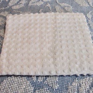 May include: A white, rectangular baby blanket with a textured, dotted surface. The blanket is made of a soft material and is placed on a blue and white patterned surface. The blanket is a light, neutral color.