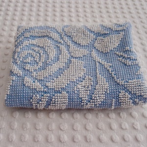 May include: A small, rectangular, light blue beaded purse with a white floral design. The purse features a repeating pattern of white beads that create a rose and leaf motif. The background is a textured white surface.