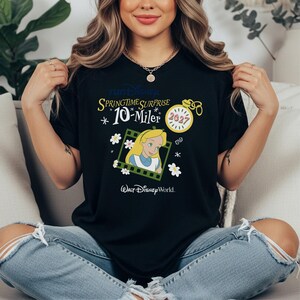 May include: A black t-shirt featuring a graphic print with the text runDisney, Springtime Surprise, 10-Miler, and Walt Disney World. The design includes an illustration of Alice, a yellow pocket watch with the year 2027, and white flower accents.
