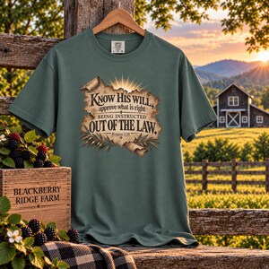 May include: A sage green short-sleeve t-shirt featuring a graphic of a tan parchment scroll with the text: Know His will, approve what is right BEING INSTRUCTED OUT OF THE LAW. Romans 2:18. The design includes sunburst rays and olive branches.