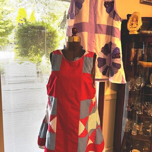 以下が含まれることがあります： エプロン風のノースリーブドレス2着。手前のドレスは赤色で、青と白の星柄パッチワークパネルが施されています。奥のドレスは白色で、紫色の縁取りと、紫色の濃淡で表現された大きな円形のフラワーモチーフの布製アップリケが特徴です。