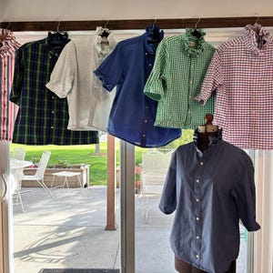 Puede incluir: Surtido de camisas en varios estampados y colores, incluyendo rayas, cuadros y vichy. Las camisas se exhiben en perchas y un maniquí. Los colores incluyen rojo, verde, azul y blanco. Las camisas tienen cuellos con volantes y botones.