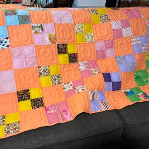 Może przedstawiać: Narzuta patchwork z powtarzającym się wzorem jednolitych brzoskwiniowych kwadratów na przemian z blokami składającymi się z czterech mniejszych kwadratów. Brzoskwiniowe kwadraty są pikowane w motyw kwiatowy. Bloki zawierają różnorodne kwiatowe i geometryczne wzory.
