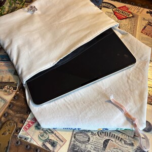 May include: A cream-coloured, padded tablet sleeve with a decorative crystal button closure. A black tablet is visible inside the open sleeve. A pink ribbon is attached. The sleeve rests on a vintage trunk.