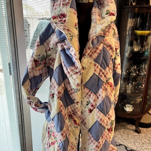 May include: A quilted coat with a hood, displayed on a mannequin. The coat features a patchwork design with blue, cream, and red plaid and floral patterns. The coat is a vintage style, with a cozy and warm appearance.