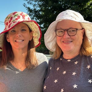 May include: A bucket hat with a white background and a red cherry pattern featuring a red brim edge, alongside a white bucket hat with a textured surface. Both hats have wide, flexible brims and are worn by two individuals in an outdoor setting.