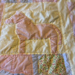 Könnte beinhalten: Nahaufnahme eines Patchwork-Quilts mit Pferdeapplikation. Der Quilt weist eine Vielzahl von Pastellfarben auf, darunter Rosa, Gelb und Blau. Das Pferd besteht aus rosa gestreiftem Stoff und ist auf einem gelben Quadrat zentriert.