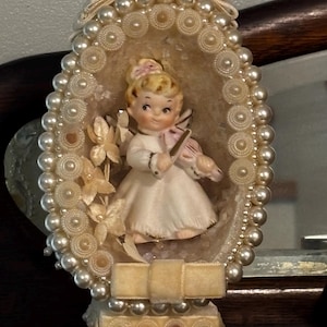 Op de afbeelding: Een decoratieve ovale display met een klein keramieken beeldje van een kind dat viool speelt. Het frame is versierd met crèmekleurige kunstparels, cirkelvormige stoffen accenten en een fluwelen strik aan de boven- en onderkant.