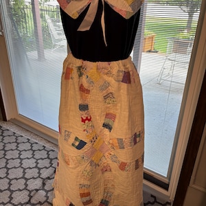 May include: A long skirt and a detachable collar made from a white quilt featuring a multicolored double wedding ring pattern. The collar is accented with a white ribbon tie at the center. The skirt has an elasticized waistband and a floor-length silhouette, showcasing intricate stitching and various floral and geometric fabric patches.