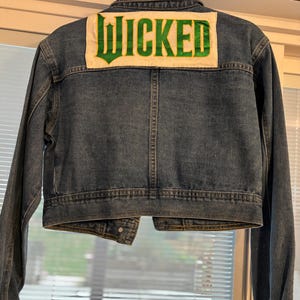 May include: A blue denim jacket with the word "WICKED" in green lettering on a white patch on the back. The jacket has long sleeves and is a medium wash.