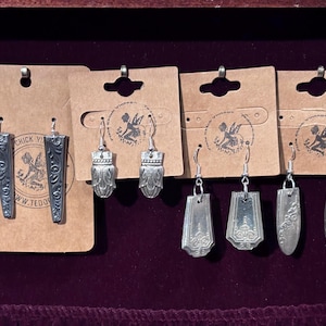 May include: A display of handcrafted silver earrings, each with a unique design. The earrings are made from repurposed silverware and hang from brown cardboard tags. The earrings are displayed against a dark purple background.