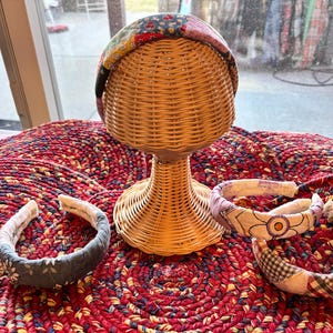 May include: A display of fabric headbands on a wicker head form. The headbands feature various patterns, including floral, patchwork, and gingham. The head form sits on a red and yellow braided rug, with a window in the background.