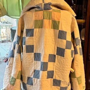 Könnte beinhalten: Gesteppte Jacke mit Kapuze, mit einem Patchwork-Design aus cremefarbenen, blauen, grünen und rosa Quadraten. Die Jacke hat eine gesteppte Textur und ein cremefarbenes Futter. Die Kapuze ist mit kleinen floralen Mustern versehen.