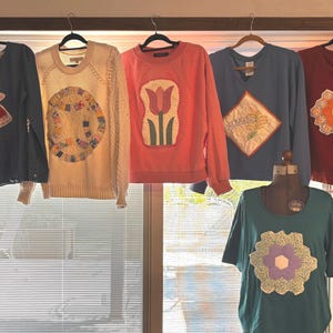 May include: A collection of six long-sleeved shirts and sweaters featuring various fabric appliqués. The garments include a blue top with a bonnet girl, a cream cable-knit sweater with a quilt ring, a coral sweatshirt with a tulip, a blue V-neck with a Nebraska diamond patch, a red top with a floral motif, and a teal shirt with a hexagonal quilt pattern.