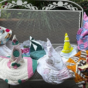 May include: A collection of six pointed hats crafted from various quilted fabrics. The hats feature diverse patterns including pink polka dots, floral prints, yellow solid fabric, and multicolored patchwork designs with wide, flexible brims.