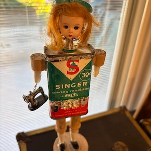 Puede incluir: Una figura de muñeca caprichosa hecha de una lata de aceite de máquina de coser Singer vintage. La figura presenta una cabeza de muñeca con cabello rojo y un lazo verde, brazos y piernas de madera y un accesorio de máquina de coser en miniatura. La lata de aceite es verde y roja.