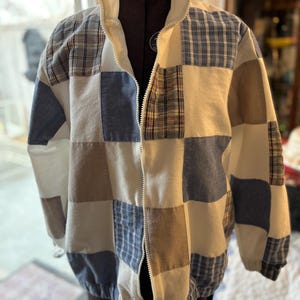 May include: A patchwork jacket with a white collar and a full zipper. The jacket features a variety of fabric squares in white, blue, and tan, with plaid and solid patterns. The jacket is displayed on a black dress form.