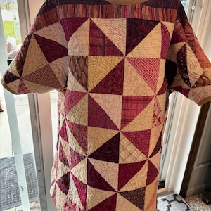 May include: A quilted tunic in shades of burgundy, cream, and gold, displayed on a mannequin. The tunic features a geometric patchwork design of triangles and squares. The neckline and sleeves are trimmed with a gold-colored fabric.
