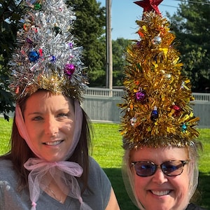 May include: Two cone-shaped hats designed to resemble Christmas trees. One hat is made of silver tinsel with a red star topper, and the other is made of gold tinsel with a red star topper. Both hats feature small colourful ornaments and pink tulle ties.