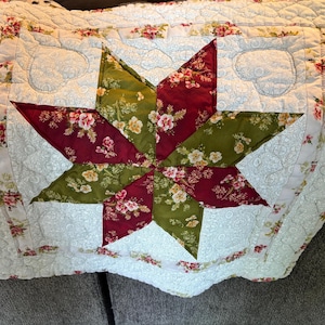 May include: A quilted throw pillow with a central star design. The star is composed of alternating sections of burgundy and olive green floral fabric. The pillow has a white background with a floral border and heart-shaped quilting.