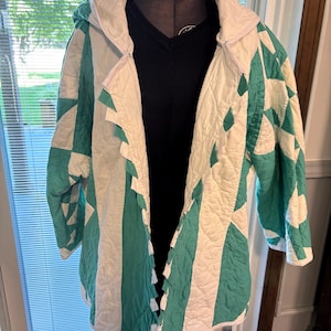 Puede incluir: Una chaqueta con capucha confeccionada en tela acolchada con un patrón geométrico en verde azulado y blanco. La prenda tiene mangas largas, una abertura frontal con un detalle dentado a lo largo de la solapa y una textura acolchada.