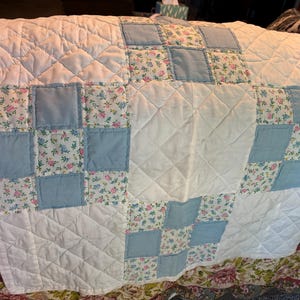 May include: A patchwork quilt featuring a repeating pattern of solid light blue squares, white squares with a diamond-stitched texture, and floral print squares with small pink and blue flowers on a white background, finished with a white border.