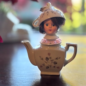 May include: A quirky teapot ornament featuring a doll's head. The cream-coloured teapot has floral patterns and gold detailing. The doll has dark hair, a pink collar, and a teacup hat, creating a unique decorative piece.