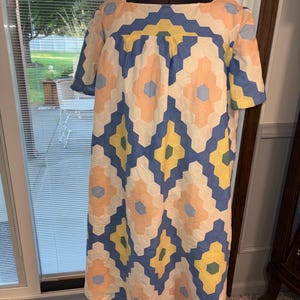 Könnte beinhalten: Ein knielanges Kleid mit einem sich wiederholenden geometrischen Muster aus ineinandergreifenden Sechsecken in den Farben Hellblau, sanftem Pfirsich, Gelb und gedämpftem Grün. Das Kleid hat kurze Ärmel, einen eckigen Ausschnitt und einen lockeren, geraden Schnitt.