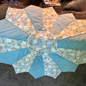 May include: A circular quilted fabric mat featuring a central floral print surrounded by alternating panels of solid light blue and a floral pattern with blue, pink, and white roses on a light background, arranged in a starburst design.