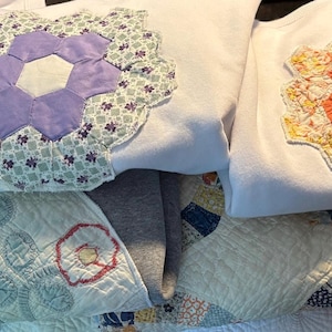 Könnte beinhalten: Eine Sammlung von Sweatshirts und Quilts. Die Sweatshirts sind weiß und lila, jedes mit einem großen, farbenfrohen Blumen-Applikation. Die Quilts sind in verschiedenen Farben und Mustern mit floralen Designs.