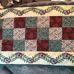 May include: A rectangular quilted table runner with a patchwork design. The centre features alternating squares of burgundy, floral, and green patterned fabric. A cream-coloured border with floral and leaf motifs surrounds the patchwork.