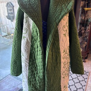May include: A long, quilted coat in shades of green and cream. The coat has a quilted pattern and embroidered floral designs on the cream panels. The coat is displayed on a mannequin, showcasing its open front and long length.