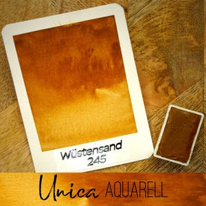May include: A watercolour paint swatch labelled "Wüstensand 245" with a rectangular shape, showing a gradient of brown hues. A small, rectangular paint pan with the same colour is next to the swatch. The text "Unica AQUARELL" is at the bottom.