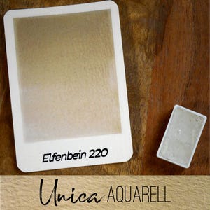 Può includere: Un campione di pittura ad acquerello beige chiaro etichettato "Elfenbein 220" e un piccolo blocco di pittura. Il testo "Unica AQUARELL" è in basso. Gli elementi sono su una superficie di legno.