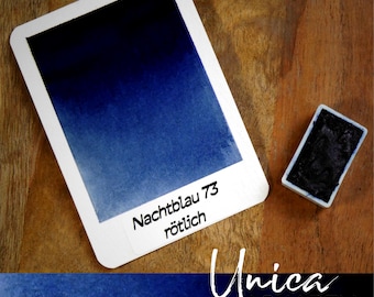 May include: A watercolor paint swatch card with a gradient of dark to light blue, labeled "Nachtblau 73 rötlich." A small, rectangular paint pan sits beside the card. The text "Unica AQUARELL" is also visible.