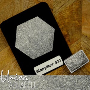 Op de afbeelding: Een zwarte kaart met een zilveren glitter zeshoek en een klein rechthoekig verfmonster. De tekst "Silberglitzer 300" staat op een wit label. De onderkant van de afbeelding heeft de tekst "Unica AQUARELL".