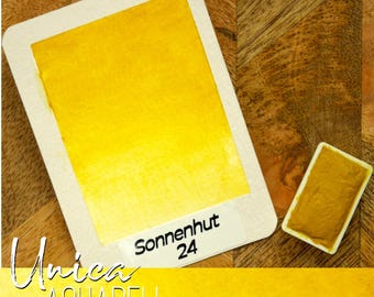 May include: A watercolor paint swatch with a vibrant yellow hue, labeled "Sonnenhut 24." The paint is on a rectangular card with rounded corners, next to a small, solid paint block. The word "Unica" and "Aquarell" are also visible.