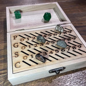 May include: A wooden box with a sliding lid, revealing a game board with numbers and letters. The game board has slots for coins, and there are five coins in the slots. The box is open and the lid is propped up.