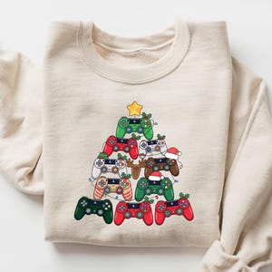 May include: Beige sweatshirt featuring a Christmas tree design made of colorful video game controllers. The controllers are red, green, white, and brown, with a gold star on top. Some controllers have festive decorations like holly and Santa hats.
